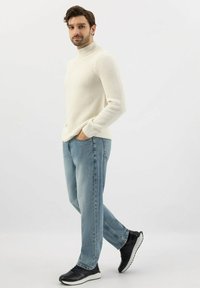 Muž stojící s rukama v kapsách, oblečený v krémovém roláku, světle modrých džínách a černých teniskách na bílém pozadí.