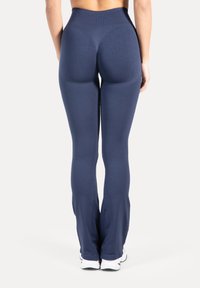 Frau trägt hoch taillierte, dunkelblaue, eng anliegende Hose mit leichtem Schlag am Saum, steht barfuß vor weißem Hintergrund.