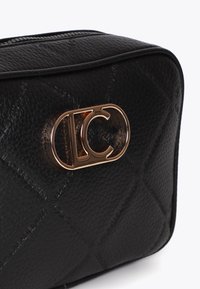 Närbild av en svart läderhandväska med ett guldtonat logoklack och texturerad yta som framhäver dess eleganta design och hantverk.