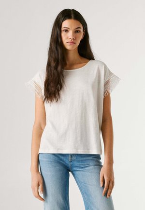 Jonge vrouw met lang donker haar, gekleed in een wit shirt met korte mouwen en kanten randje, en lichtblauwe spijkerbroek, staand tegen een effen achtergrond.