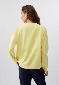 Femme aux longs cheveux portant un sweat-shirt jaune pâle et un pantalon foncé, vue de dos sur un fond clair et uni.