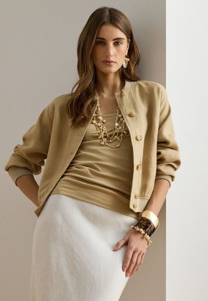 Γυναίκα με μπεζ σακάκι, ανοιχτό καφέ τοπ, λευκή φούστα, στρωματοποιημένο κολιέ από κοχύλια, μεγάλο βραχιόλι και σκουλαρίκια από κοχύλια, πόζα μπροστά σε ουδέτερο φόντο.