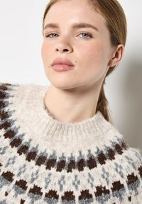 Sweter robiony na drutach z wysokim, prążkowanym kołnierzem. Kremowa baza z geometrycznym wzorem w brązie i szarości. Miękki, teksturowany materiał wełniany.