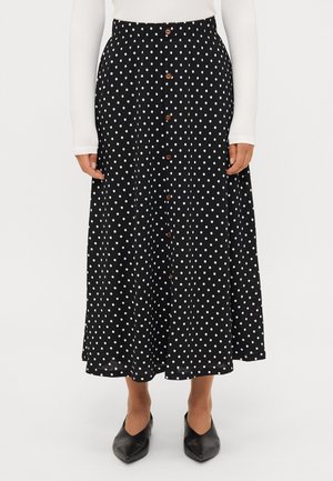 Jupe midi noire à pois blancs, boutons sur le devant, portée avec des chaussures noires pointues et un haut blanc à manches longues.