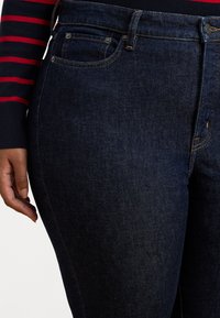 Mørke denimjeans med en glatt tekstur, en klassisk fem-lommers design og kontrasterende sømmer. Nærbilde som viser midje og lommer.
