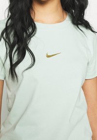 Ljusgrön bomullströja med rund halsringning, prydd med en guld Nike-swooshlogga på bröstet. Korta ärmar, mjuk textur.