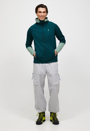 Glunderende man die een teal- en lichtgroene jas met rits draagt, lichtgrijze cargobroek en zwarte en gele wandelschoenen, handen in zijn zakken.