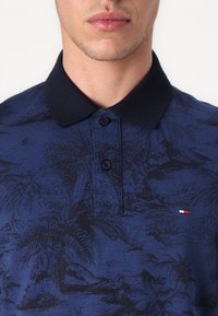 Granatowa koszulka polo wykonana z teksturowanej tkaniny z wzorem palm, dopełniona jednokolorowym czarnym kołnierzykiem i plisą z trzema guzikami.