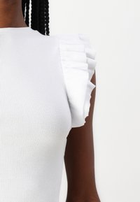 Top blanco de canalé con mangas cortas con volantes y un diseño ajustado, que resalta la textura y los acentos estructurados.