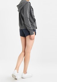 Sudadera gris con capucha y logo impreso, combinada con shorts oscuros y zapatillas blancas. Tejido suave y corte relajado, mostrada de lado.