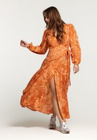 Oranje wikkeljurk met bloemenpatroon, lange opgeblazen mouwen en een zijslit. Gepaard met zilveren sport sneakers.