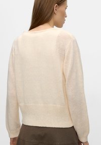 Jasno beżowy sweter z dzianiny o luźnym kroju, żebrowanym wykończeniu i odkrytych plecach. Materiał wydaje się miękki i ma lekko przezroczystą teksturę.