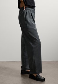 Pantaloni grigi su misura con una vestibilità rilassata, caratterizzati da pieghe frontali, tasche laterali e una texture liscia. Abbinati a mocassini neri.