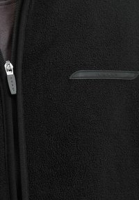 Giacca in pile nera con zip frontale, tessuto testurizzato e una piccola patch del logo nera vicino alla spalla per il branding.