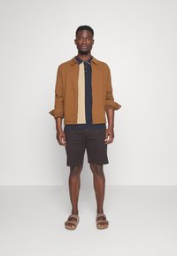 Bruine, gestructureerde jas over een beige en marineblauwe polo, gecombineerd met donkere shorts en bruine sandalen. Effen achtergrond benadrukt de outfit.