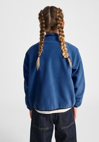 Fleecejack in blauw, met een hoge kraag en lange mouwen. De gestructureerde stof heeft een zachte, pluizige uitstraling. Gedragen over een donkere spijkerbroek.