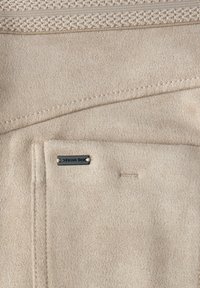 Beige mockasinfärgade tyger med en strukturerad yta, med en sydd design, en ficka med en liten metalltagg och en slät kant.