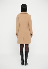 Abrigo de lana marrón claro con una silueta entallada, cuello con muesca y tres botones negros en las mangas; combinado con botas negras hasta la rodilla.