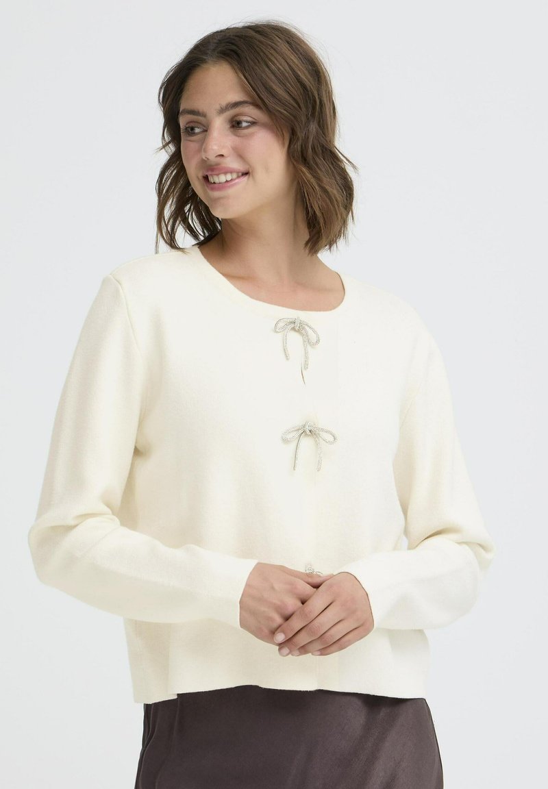 Ivory pulover iz pletenine z okroglim izrezom in dvema dekorativnima srebrnima pentljama spredaj. Dolgi rokavi in skrajšana oblika. Gladka tekstura.