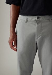 Pantalon gris clair avec une finition texturée, doté d'une fermeture à bouton et de poches latérales. Le tissu semble durable et structuré.