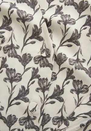 Cremefarbener Stoff mit schwarzem Blumenmuster, versehen mit stoffüberzogenen Knöpfen und sichtbaren Nähten in einem gefalteten Abschnitt.