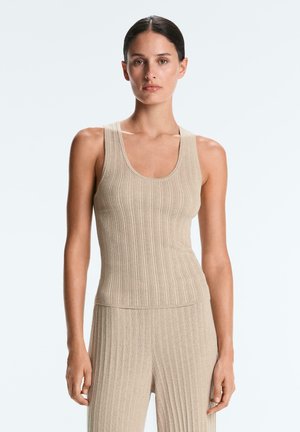 Nő áll előre nézve, bézs színű bordás ujjatlan felsőt és hozzáillő bordás nadrágot viselve, egyszínű világos háttér előtt.