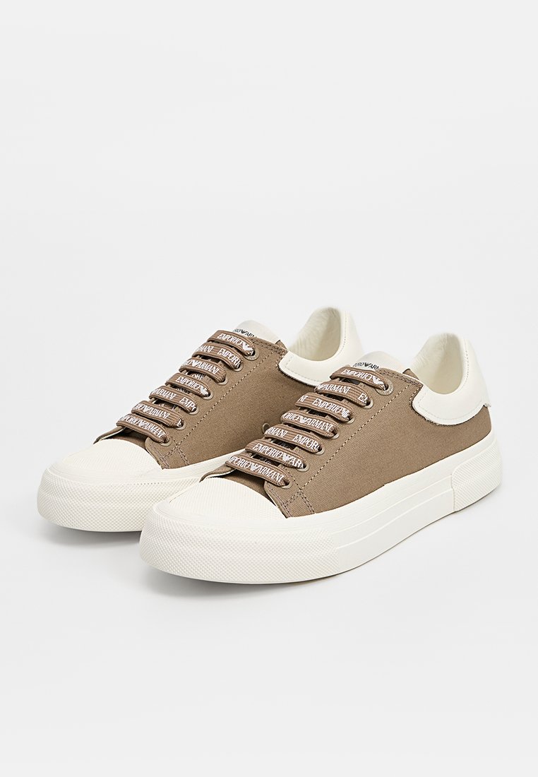 Braune Canvas-Sneaker mit einer weißen Gummisohle, die auffällige Schnürsenkel und Logo-Akzente auf der Zunge aufweisen. Glatte Textur und abgerundetes Zehen-Design.