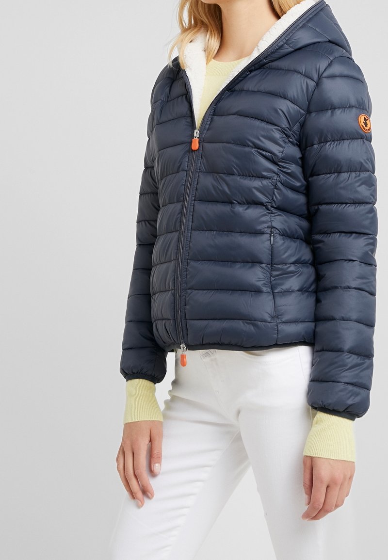 Femme portant une doudoune matelassée bleu marine avec capuche et tirettes de fermeture éclair orange, associée à un pantalon blanc et un pull jaune clair.