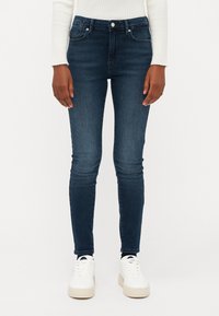 Σκούρες μπλε στενές τζιν με ψηλή μέση, πέντε τσέπες και διακριτική φθορά. Συνδυασμένες με λευκά sneakers σε απλή φόντο.