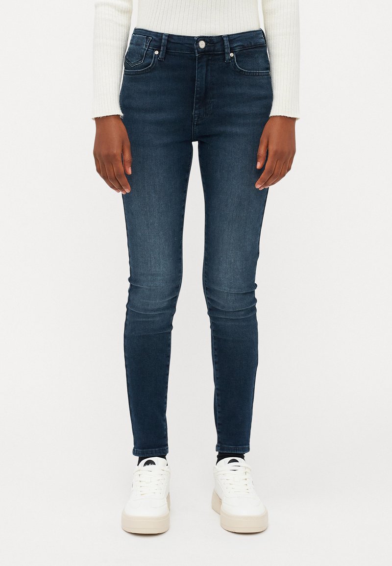 Σκούρες μπλε στενές τζιν μ�ε ψηλή μέση, πέντε τσέπες και διακριτική φθορά. Συνδυασμένες με λευκά sneakers σε απλή φόντο.