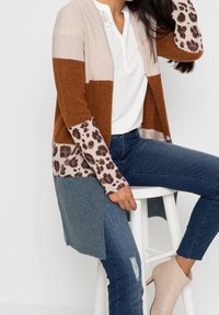 Färgglad cardigan med beige, brun och blå sektioner, med leopardmönstrade detaljer på ärmarna. Mjuk stickad material, öppen framsida.
