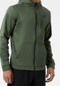 Sudadera con capucha de cremallera en verde oliva, hecha de tela suave, con capucha, bolsillos frontales y un logo negro en el pecho.