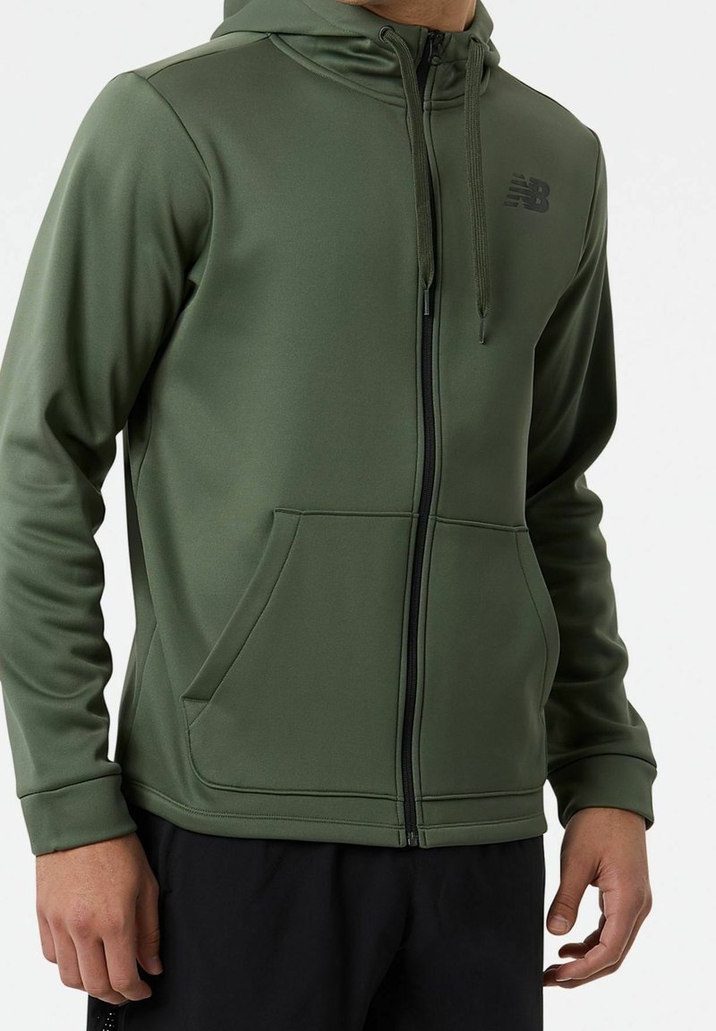 Sudadera con capucha de cremallera en verde oliva, hecha de tela suave, con capucha, bolsillos frontales y un logo negro en el pecho.
