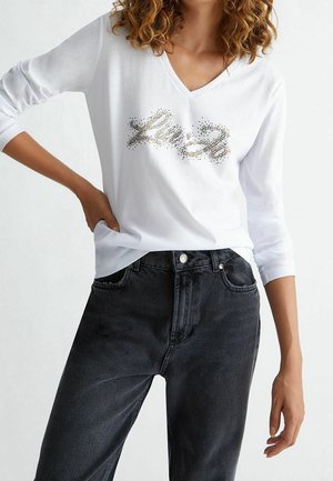 White long-sleeve V-neck shirt with a shimmering gold design on the front, paired with high-waisted dark denim jeans.