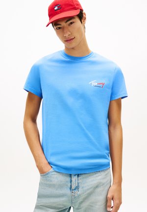 Joven con gorra roja de Tommy Jeans, camiseta azul claro de Tommy Jeans y vaqueros azul claro, de pie con una mano en el bolsillo.