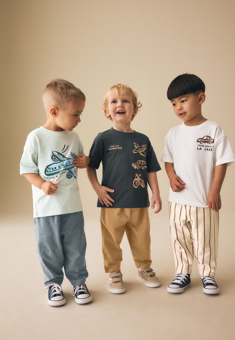 Trois jeunes garçons debout ensemble portant des t-shirts graphiques et des pantalons décontractés, en interaction dans un studio au décor neutre.