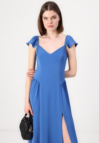 Robe bleue avec des manches courtes à volants, décolleté en V, corsage ajusté et jupe fluide avec fentes sur les côtés, accompagnée d'un sac à main noir.