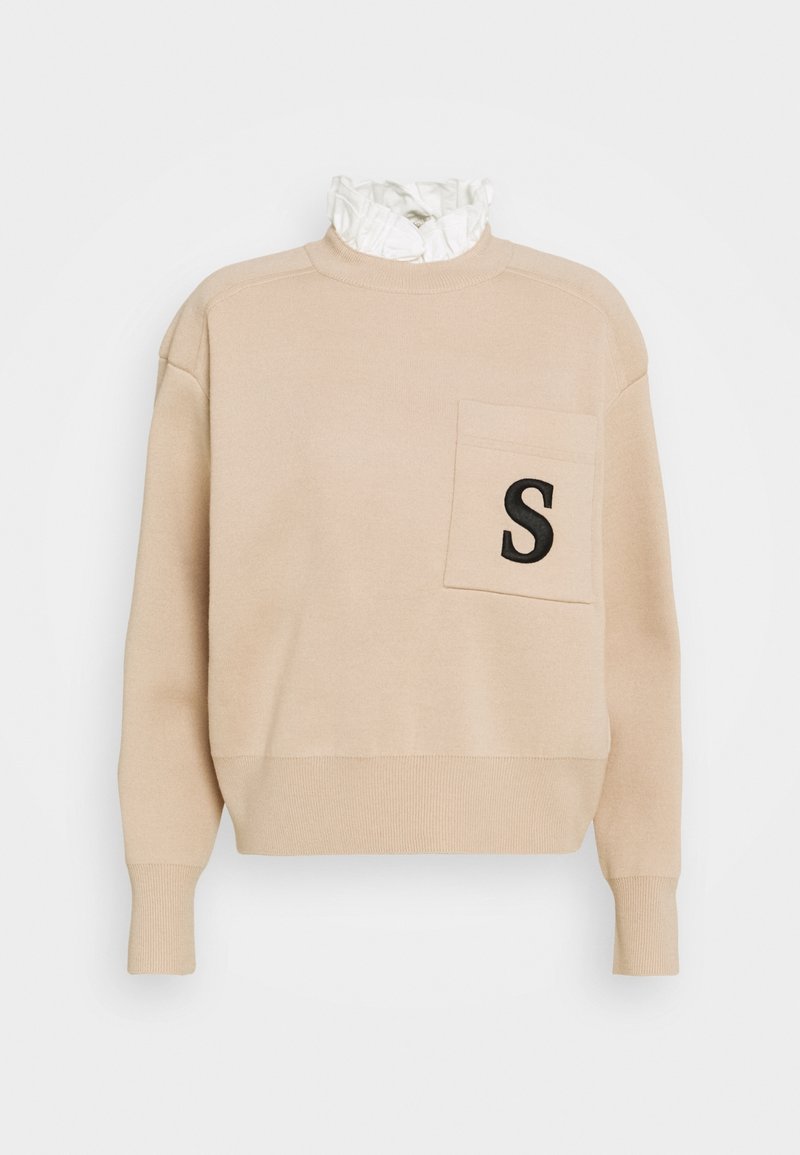 Pull beige à manches longues avec poignets côtelés, une poche poitrine ornée d'un "S" noir, et un détail de col blanc à volants.