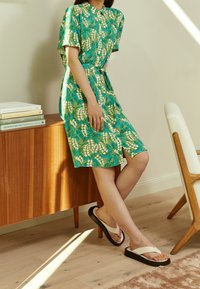 Grün und blau gemustertes Kleid mit durchgehender Knopfleiste, kurzen Ärmeln und Taillenknoten. Schwarz-weiße Flip-Flops, Holzmöbel im Hintergrund.