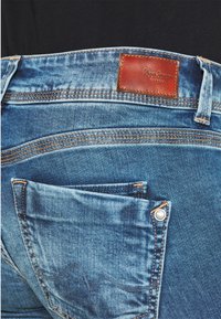 Zbliżenie na tylnią kieszeń niebieskich dżinsów, z brązową skórzaną naszywką marki Pepe Jeans oraz pomarańczowymi detalami stębnowania.