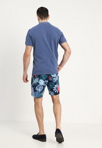 Blå polo med korte ærmer, parret med mørk badebukser med blomsterprint, der har grønne blade og farverige blomster.