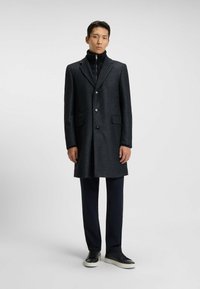 Cappotto lungo grigio scuro con texture, colletto a scienziato, chiusura a bottoni e due tasche anteriori. Indossato sopra un dolcevita nero e pantaloni blu navy.