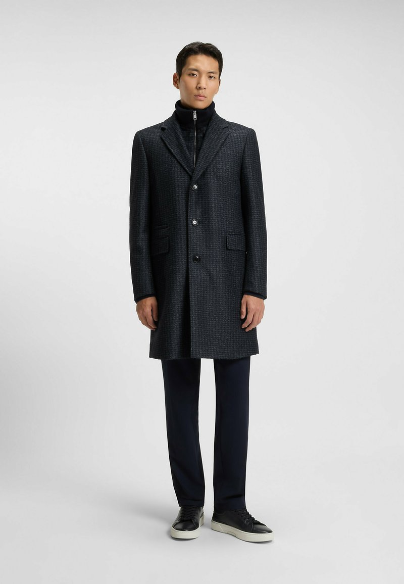 Cappotto lungo grigio scuro con texture, colletto a scienziato, chiusura a bottoni e due tasche anteriori. Indossato sopra un dolcevita nero e pantaloni blu navy.