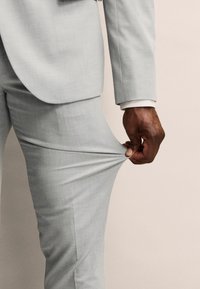 Traje de color gris claro compuesto por una tela texturizada; una mano agarra la pierna del pantalón, mostrando su ajuste a medida y acabado suave.