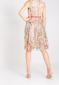 Blommönstrad broderad klänning i mjuk rosa tyg, med en åtsittande överdel, utställd kjol och ett rött band i midjan. Bärs tillsammans med nude-hälar.