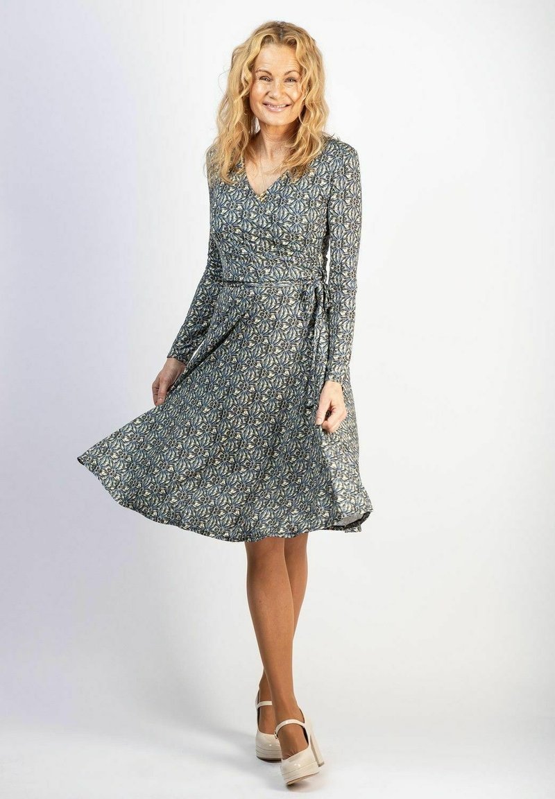 Langsleeve wikkeljurk in een blauw en beige bloemenprint, met een fitted taille en een uitlopende rok, gecombineerd met lichtgekleurde platform schoenen.