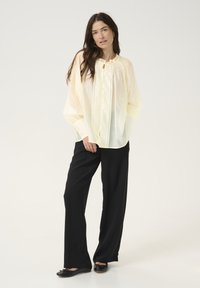 Camicia button-up leggera di un giallo pallido con scollatura arricciata, abbinata a pantaloni neri a gamba larga e scarpe nere ballerine. Tessuto liscio e morbido.