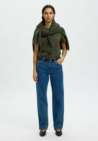 Olijfgroen t-shirt, oversized groene trui over de schouders gedrapeerd, blauwe wijde jeans, zwarte riem en zwarte puntige schoenen.