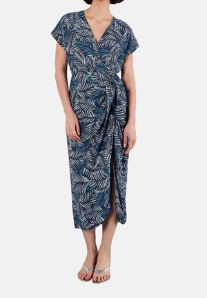 Woman wearing a blue wrap dress with white and teal leaf prints, short sleeves, and a front slit, paired with silver sandals.