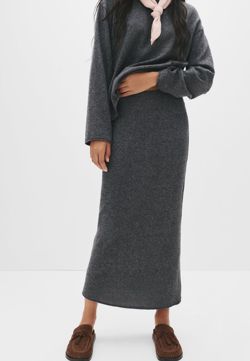 Gonna grigia, strutturata, fino al ginocchio, abbinata a un maglione oversized coordinato e ciabatte in stile mocassino marroni. Sciarpa rosa intorno al collo.